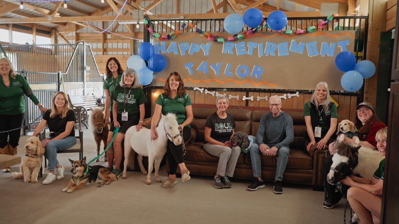 Taylor, her pet parents, and attendees at her retirement party pose around a banner that reads "Happy Retirement, Taylor"