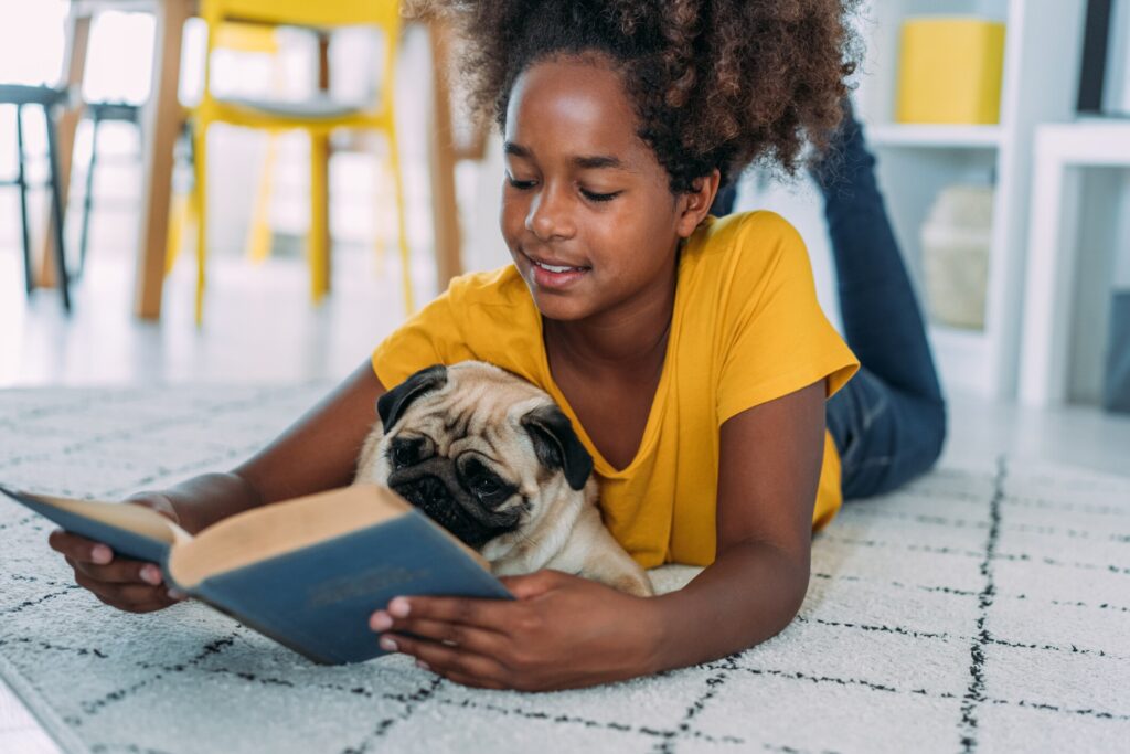 little girl reads book to pug while lying on floor
