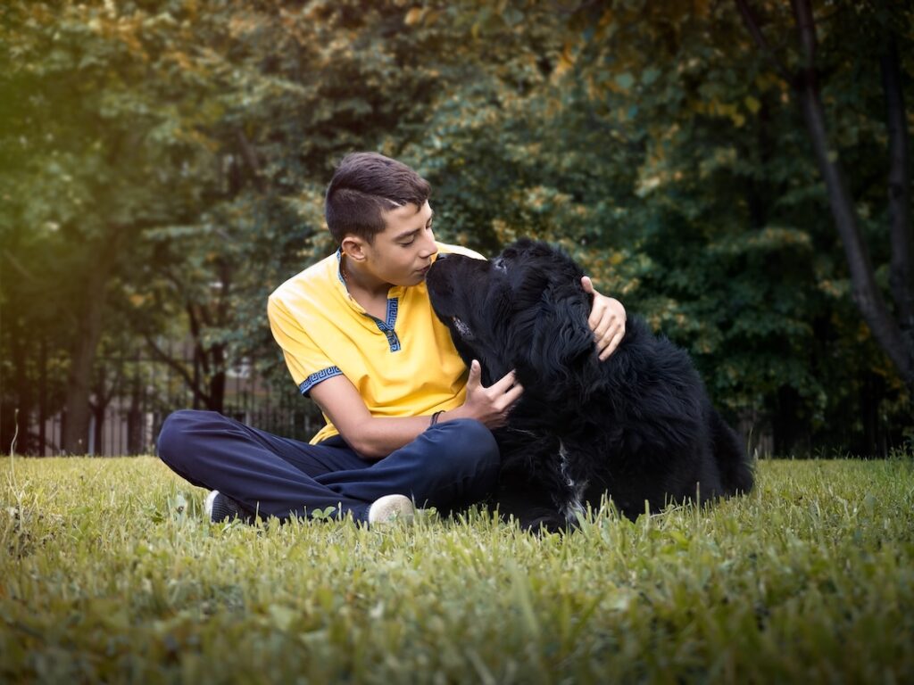 Young man sitting on grass and affectionately embracing a black Newfoundland dog in a park.