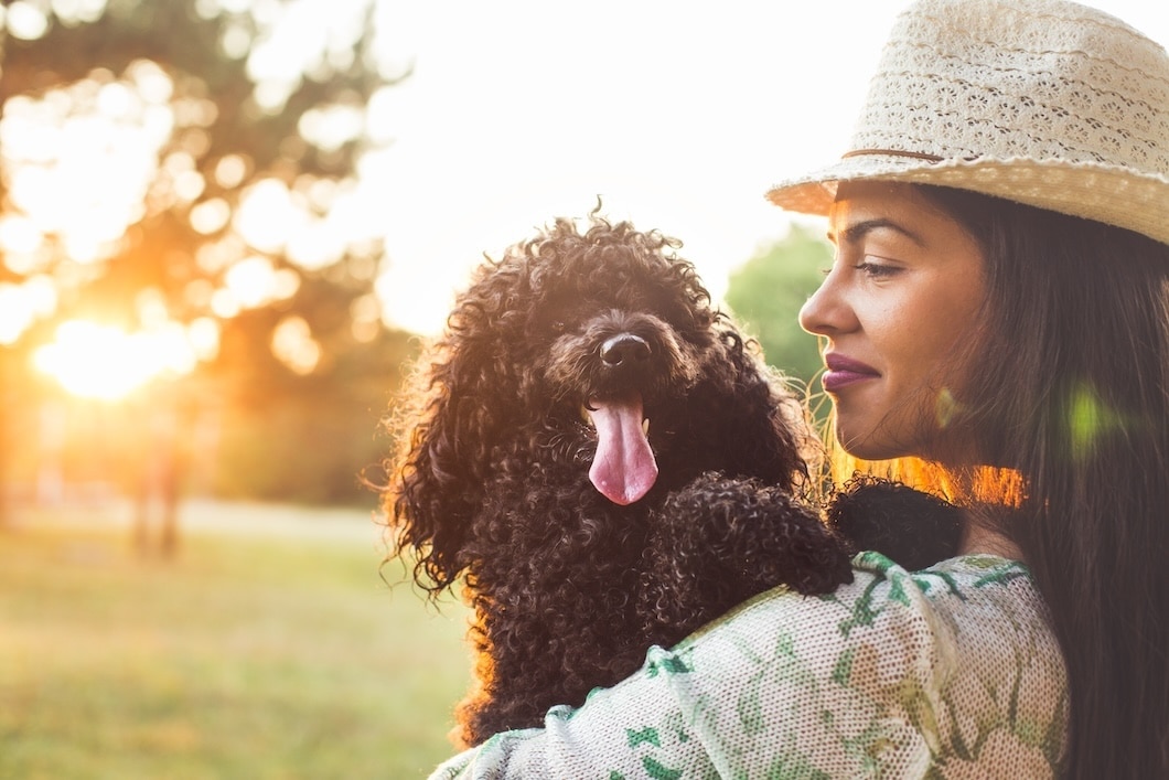 Woman wearing a sun hat holding a black curly-haired dog outdoors at sunset.